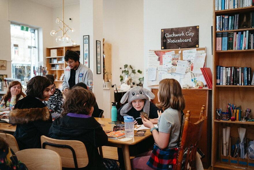 Children seated around a work table in a large studio space, interacting with one another and writing in notepads, a wall of bookshelves behind them.