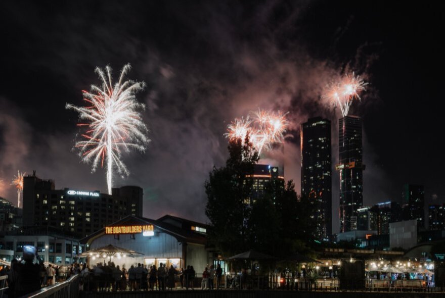A fireworks display over city buildings at night, with the Boatbuilder's Yard in the foreground, and people standing around outside.
