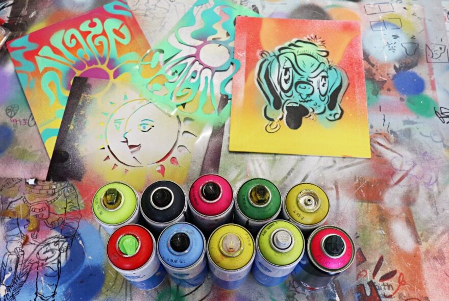 Two rows of coloured paint spray cans and a selection of street art stencils, on a flat work surface.