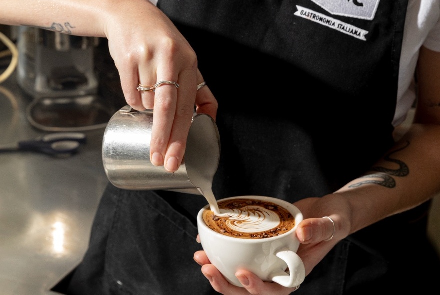 A barista in a black apron pouring milk from a frothing jug into a cappuccino.