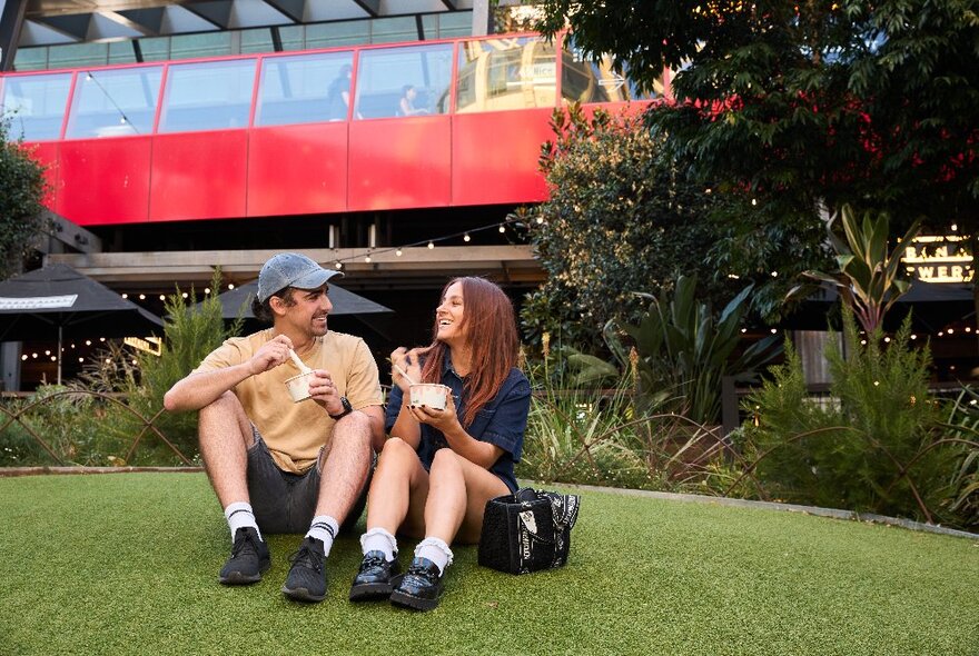 A couple enjoying ice cream from paper cups on a grassy area in front of a red and glass building.