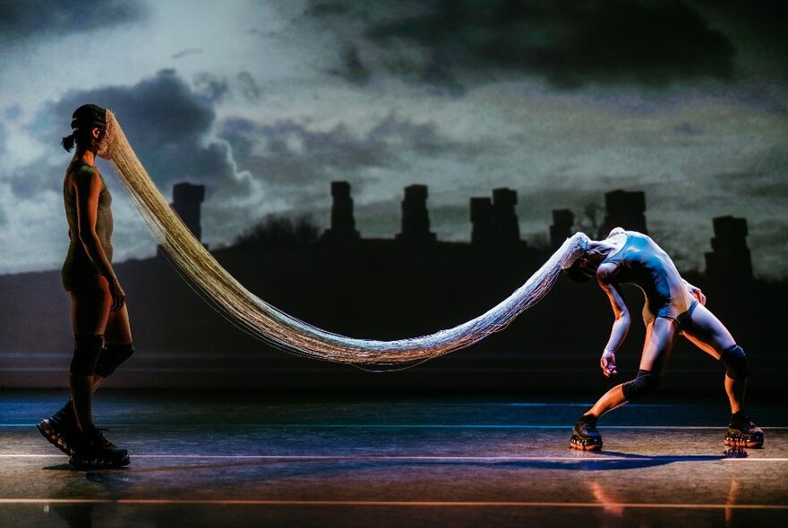 Two members of dance group Neon Dance performing on a darkened stage, their faces joined by many fine threads, a background that resembles the outline of Stonehenge. 