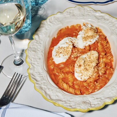 A plate of tomato fusilli with three slices of mozzarella on top in a delicately patterned white plate, alongside a glass of white wine.