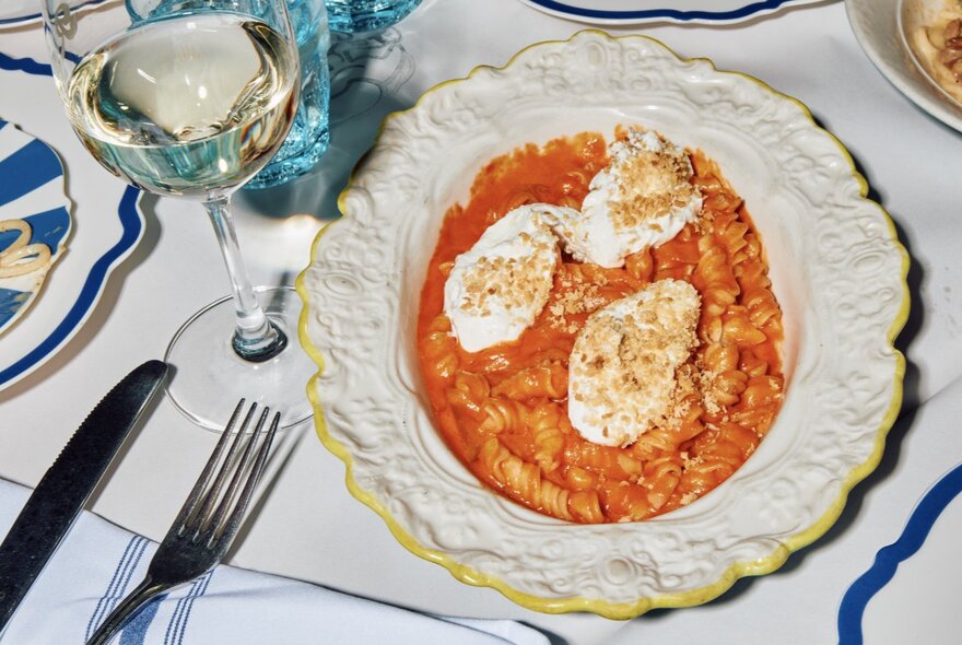 A plate of tomato fusilli with three slices of mozzarella on top in a delicately patterned white plate, alongside a glass of white wine.