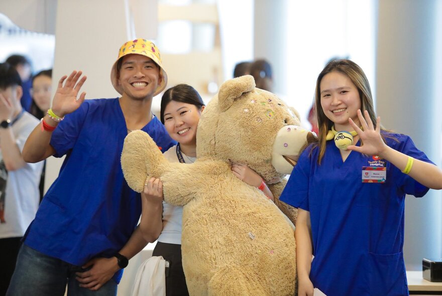 Three adults smiling and waving while holding up a life-sized soft toy teddy bear, at the Kids Day Out.