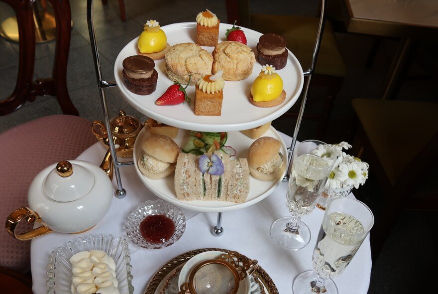 A tiered platter of High Tea treats including cakes, sandwiches and mini rolls, with champagne glasses and a teapot alongside it.