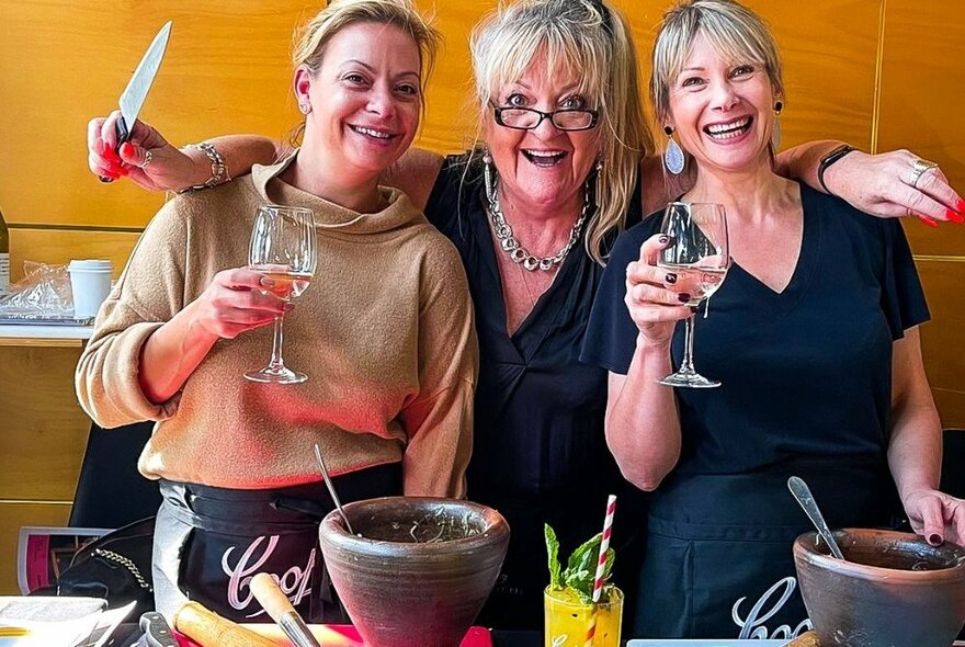 Three women at a Thai cooking workshop with wine glasses, and one holding a chef's knife.