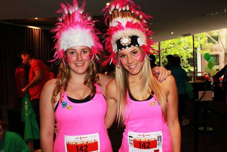 Two women wearing pink singlets with racing numbers and elaborate headdresses. 