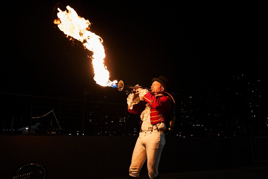A performer blowing fire through a trumpet shaped instrument, against a black night sky, as part of the Firelight Festival.