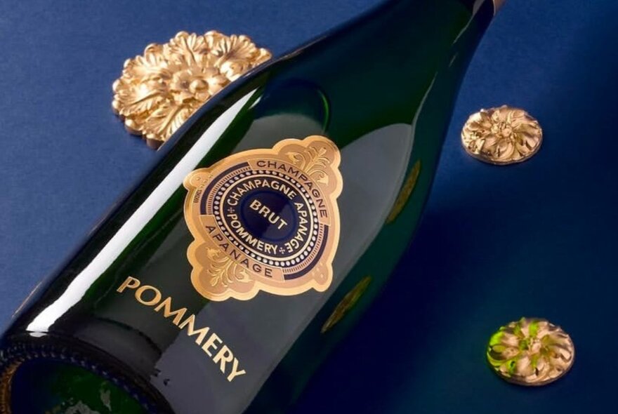 A champagne bottle with gold insignia and gold lettering, resting on its side on a dark blue surface, with gold seals.