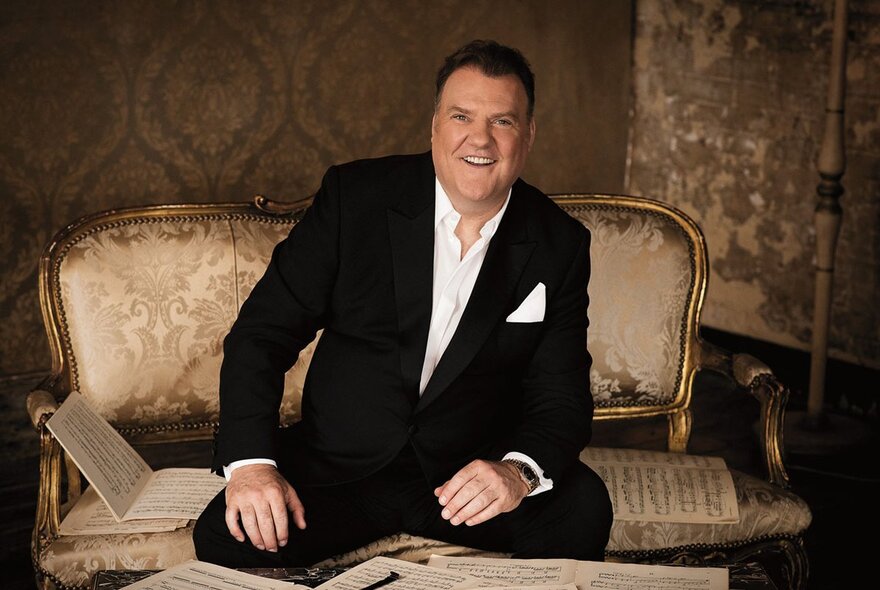 Welsh baritone Sir Bryn Terfel sitting on a vintage couch surrounded by sheet music, smiling at the camera.