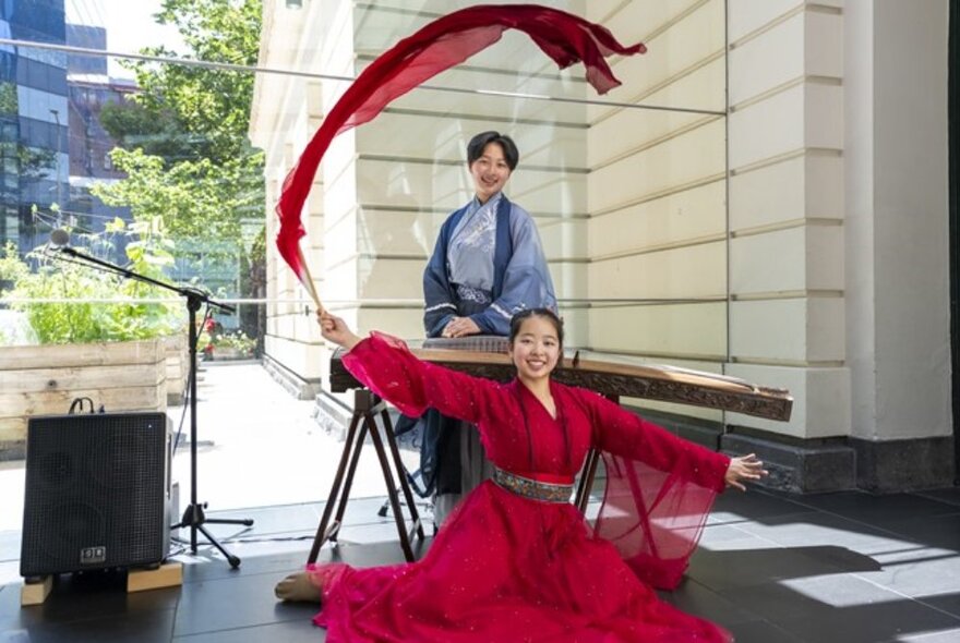 Traditional dancer with a red dress and ribbon curling in the air, another woman looking on. 