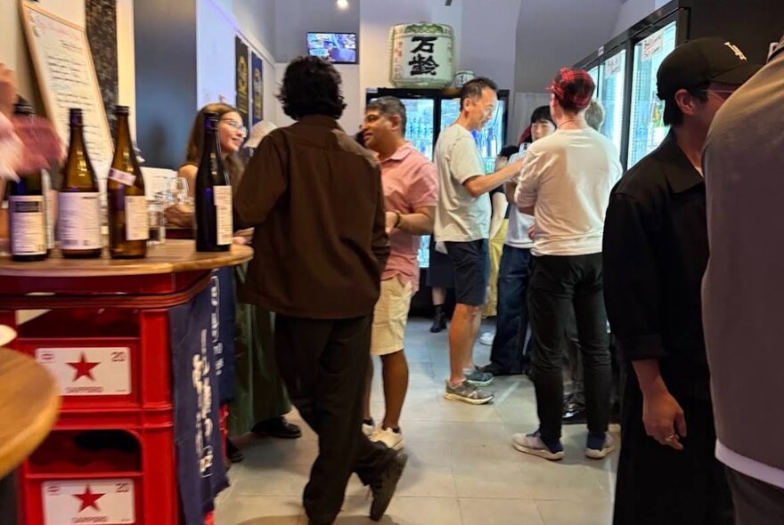 People standing with drinks in a Japanese bottle shop/bar in front of fridges.