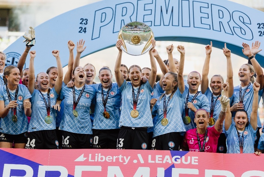 A women's soccer team victoriously holding up a premiership trophy and posing for a group shot.