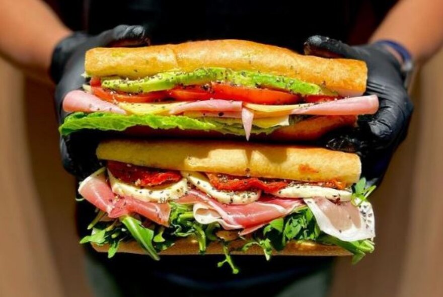 A pair of delicious looking filled french baguettes being held one above the other by two hands in black gloves.