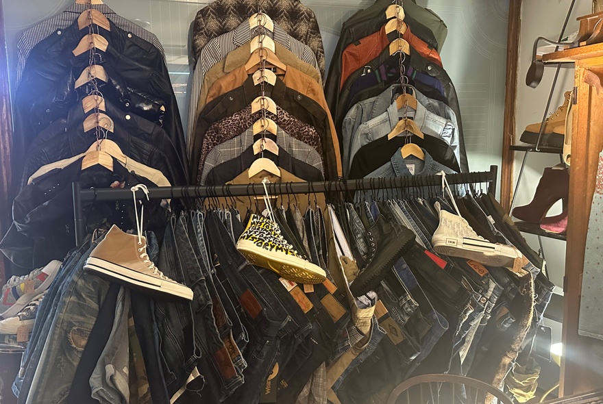 A rack of assorted casual apparel, including folded flannel or patterned shirts on hangers, jackets, and denim trousers with high-top sneakers displayed on top.