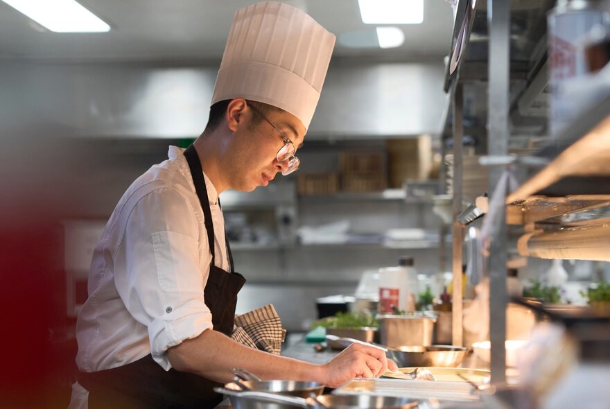 Chef wearing a chef's hat, apron and spectacles, working in a stainless steel restaurant kitchen.