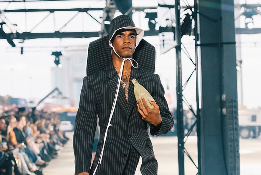 A male fashion model walking a fashion runway in an outdoor undercover space.