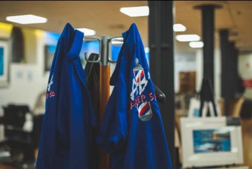 Branded barber coats hanging on a hatstand in a room with black pillars and overhead lights.