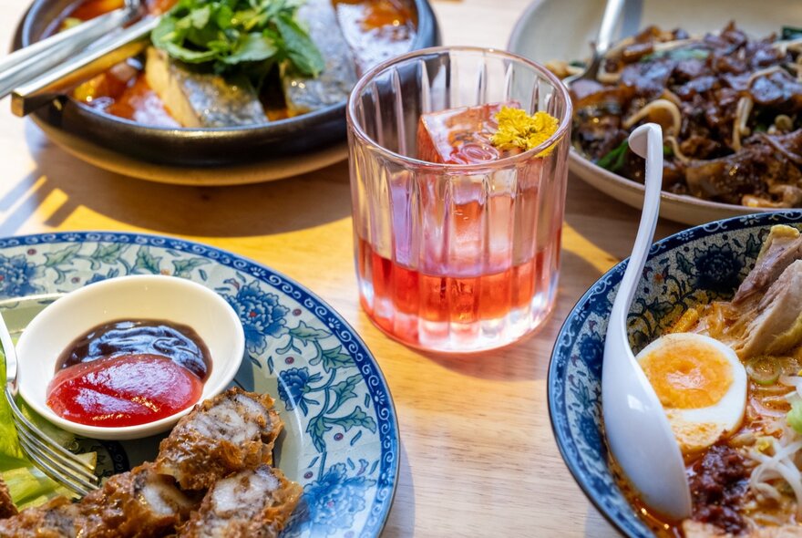 Various plates of Malaysian food and a Campari based cocktail in the middle. 