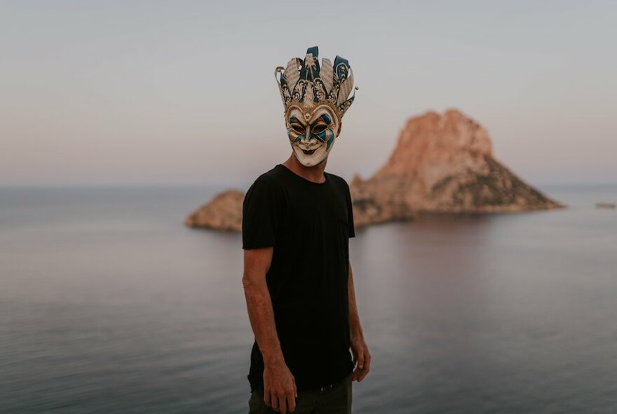 Person wearing black pants and a black t-shirt and an elaborate Venetian carnival style full face mask, with the sea and a rocky outcrop behind them.