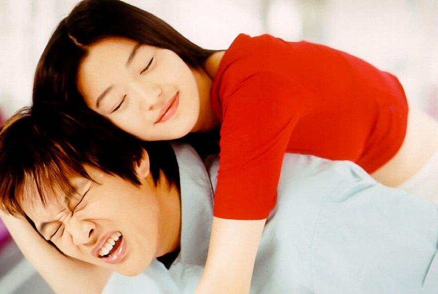 A Korean woman in a red t-shirt jumping on the back of her Korean boyfriend, who is grimacing with effort.