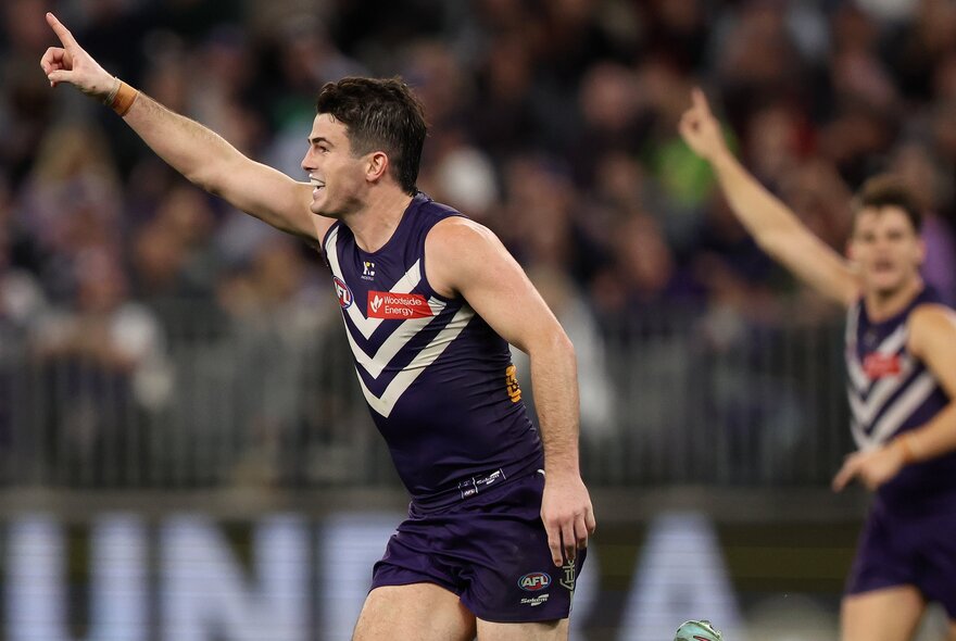 Two Fremantle AFL players on a sports ground cheering with the blurred fans in the stadium around them.
