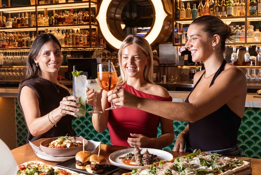 Three friends smiling and raising their glasses above a table laden with platters of food, at Oche Melbourne.