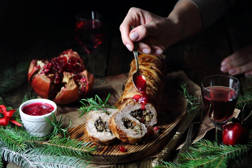 Hand pouring cranberry sauce onto stuffed turkey roll on a wooden platter surrounded by holly, pomegranate and glass of wine.