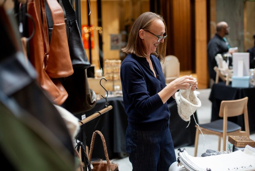 A woman standing smiling and knitting at a craft market.