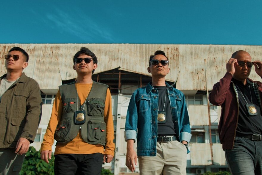 Four men walking together in a line, looking towards the camera, all wearing sunglasses and police ID pendants; a still from a movie.