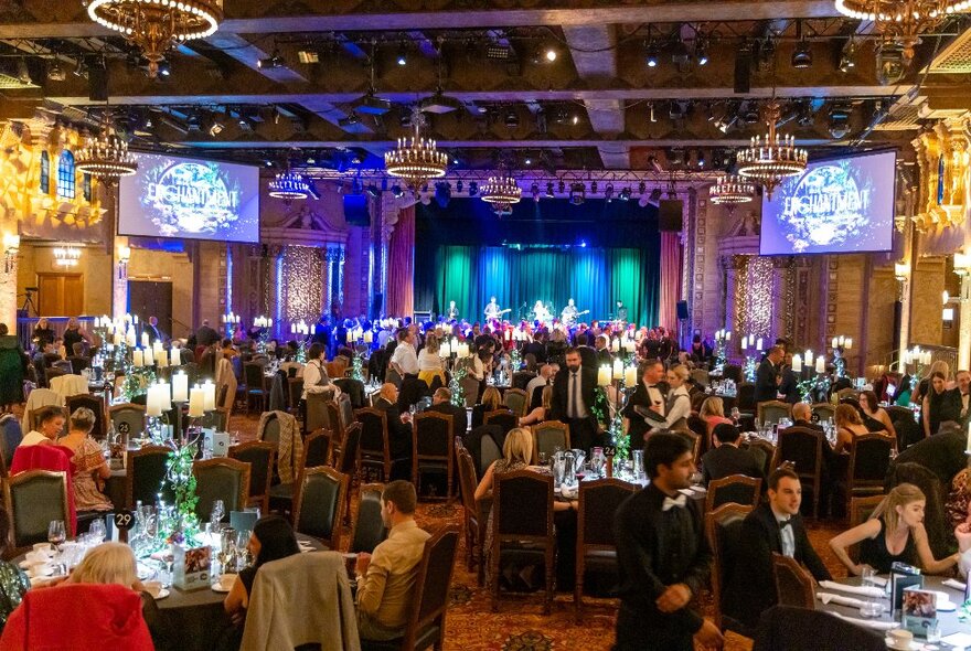 Crowded dining event with tables of diners wearing formal evening wear seated on a dance floor with band playing live on stage.