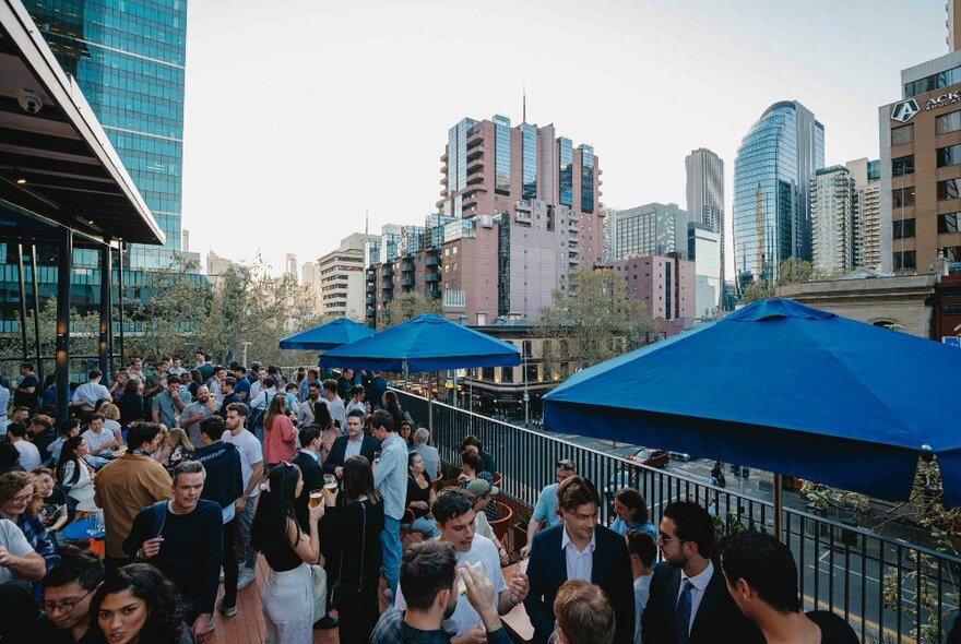 People outside under shade umbrellas at a crowded rooftop bar.