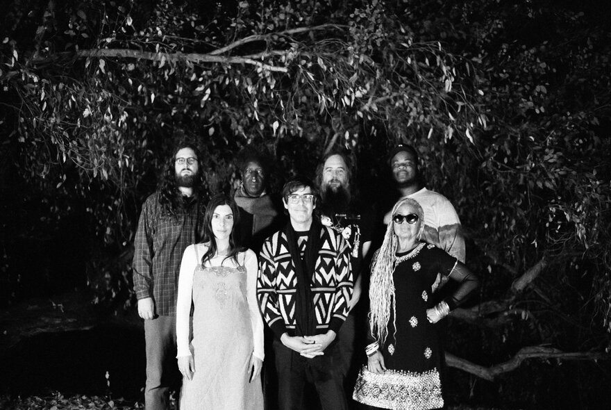 Black and white image of seven musicians, including Saul Williams and Carlos Niño, standing outdoors at night among trees.