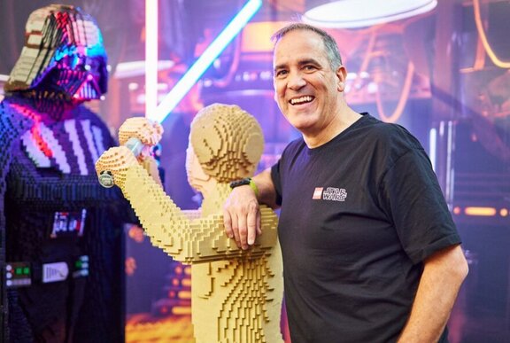 'Brickman' Ryan McNaught, smiling  nas he leans on a LEGO sculpture of Luke Skywalker who is clashing light sabers with Darth Vader.