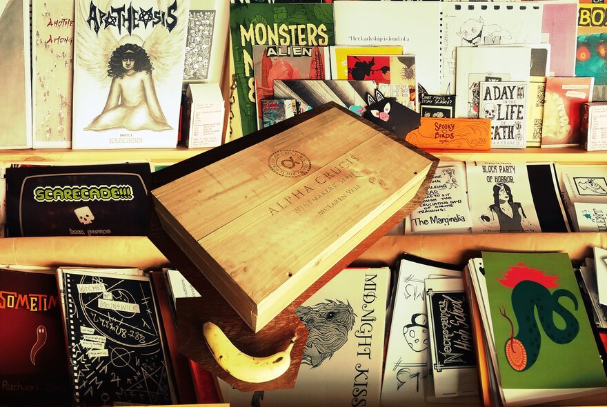 A light brown, rectangular wooden box sitting tilted on a shelf filled with various books, zines, and comics.