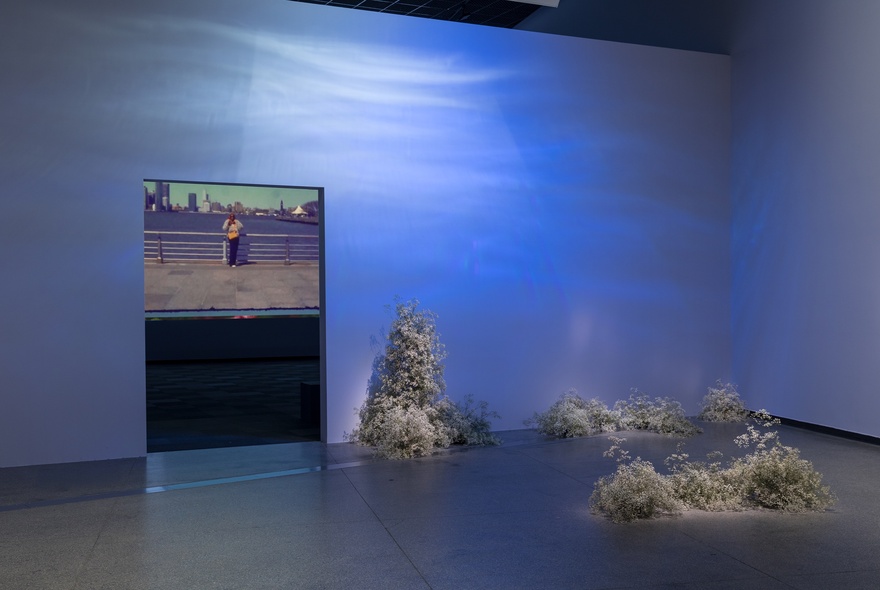 A gallery wall with a digital projection of water, tufts of leaf matter on the floor and a digital image of a person on a bridge seen through a doorway.