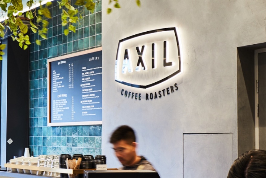 Person making coffee behind a just visible coffee machine, behind them is a blackboard menu on a green tiled wall and a large logo that reads AXIL COFFEE ROASTERS.