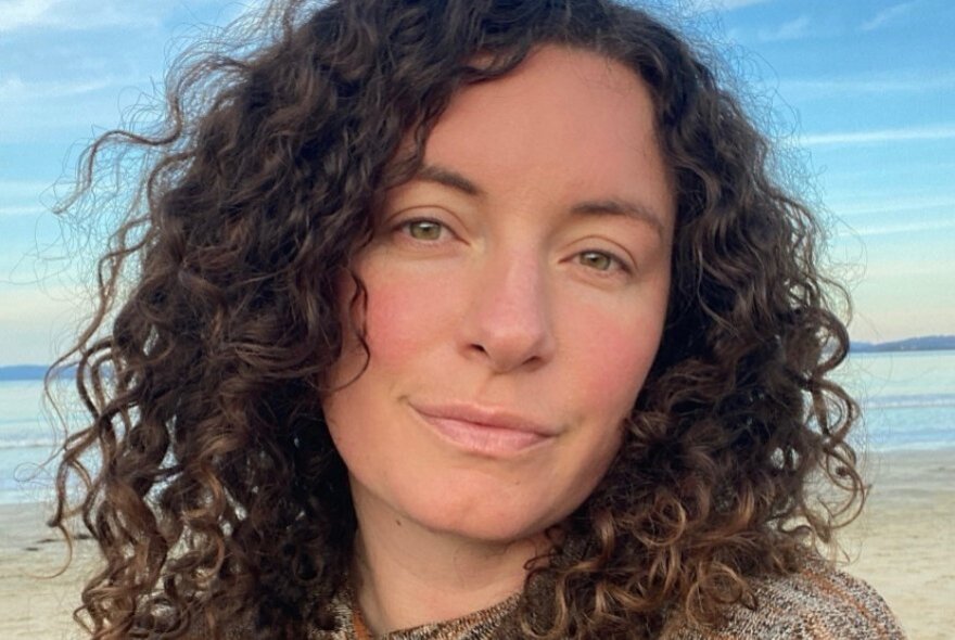 Poet Chloe Mayne, with curly brown hair framing their face, a neutral expression, posing outdoors with sand and sea behind them.
