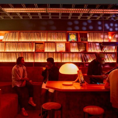 Vinyl cocktail bar with walls lined with shelves of LPs, people seated on bench seating with atmospheric lamp lighting.
