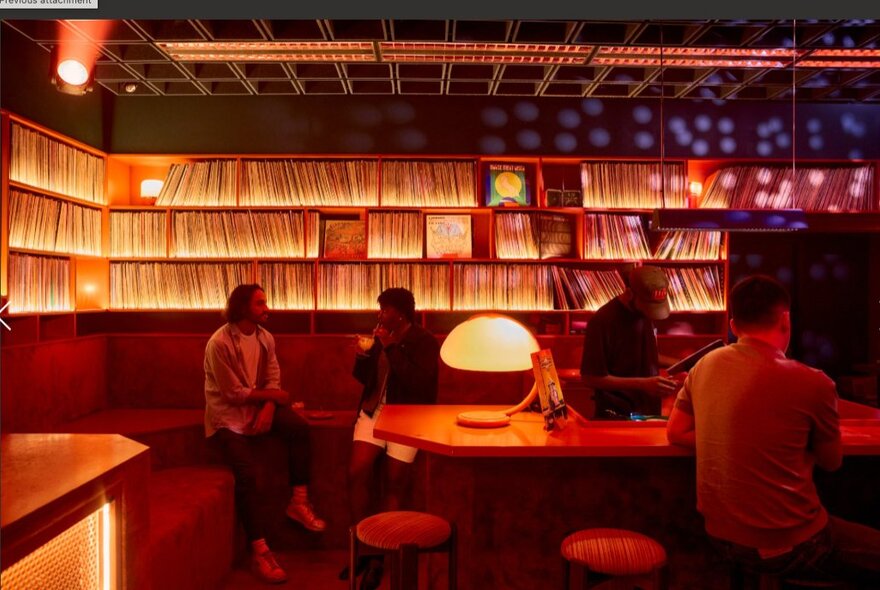 Vinyl cocktail bar with walls lined with shelves of LPs, people seated on bench seating with atmospheric lamp lighting.