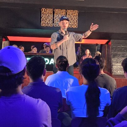Comedian holding a microphone with arm outstretched, performing on a stage in front of seated audience under purple lighting.
