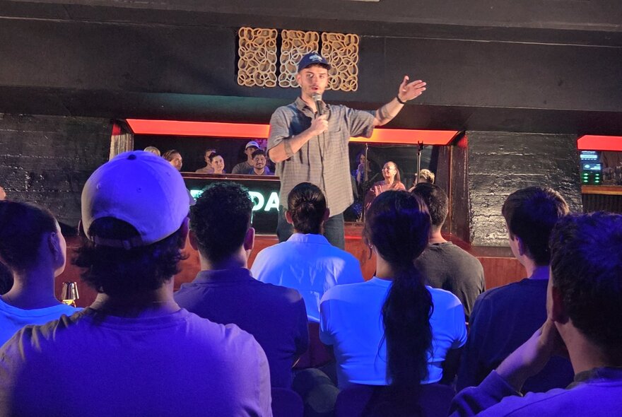 Comedian holding a microphone with arm outstretched, performing on a stage in front of seated audience under purple lighting.