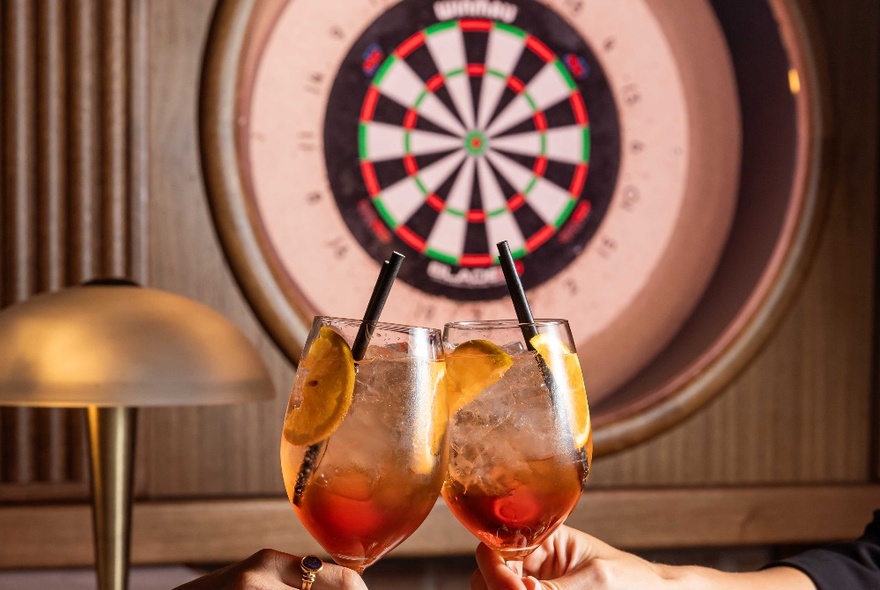 Two hands bringing together two cocktails in glasses with a dart board in the background.