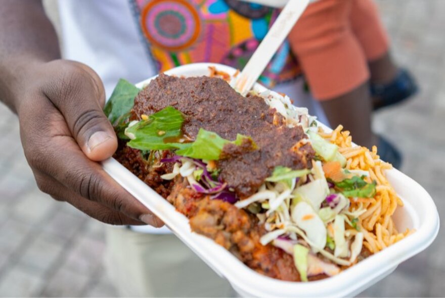 A hand offering a takeaway plate of African food with a fork sticking out. 