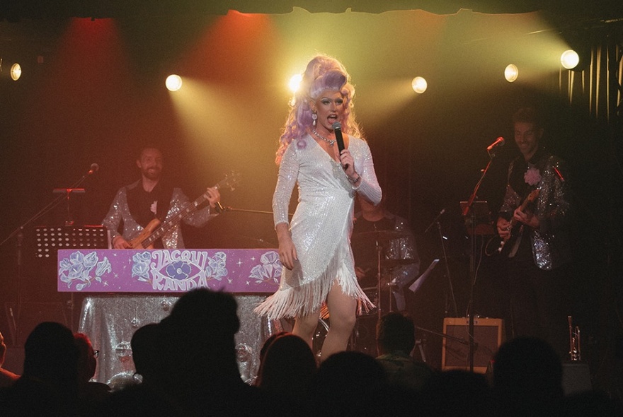 A drag queen with big hair performing on stage with a live band in the background. 