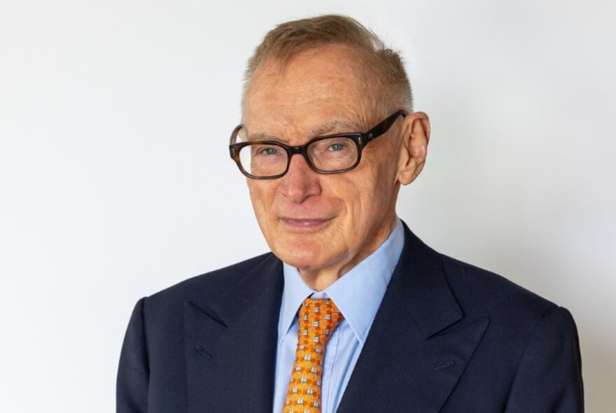 Portrait of former NSW Premier, Bob Carr, wearing a blue suit and orange tie, smiling.