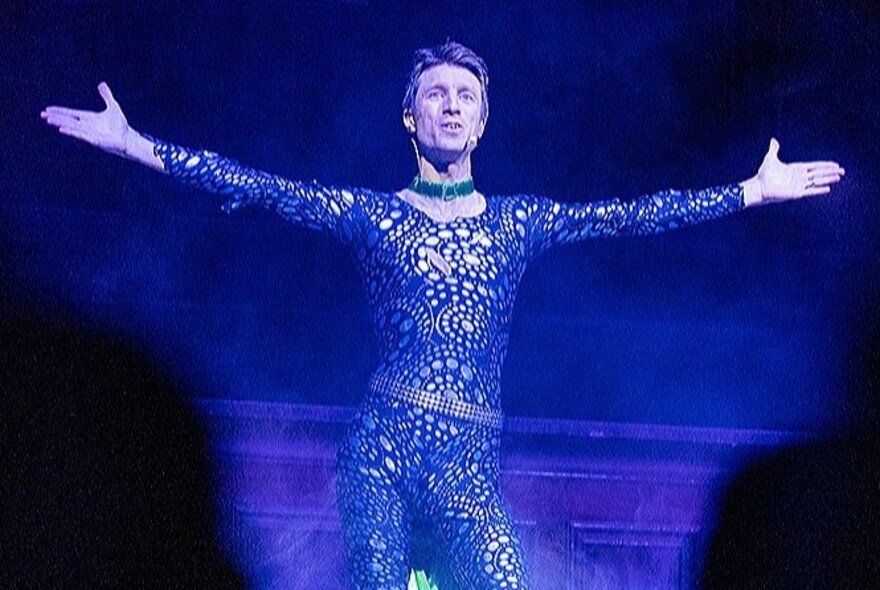 Comedian Sammy J, on stage in a skin-tight lace jumpsuit with arms spread wide.