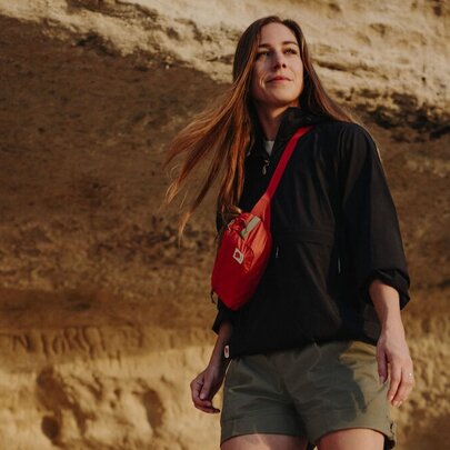 A woman in front of a natural rock formation wearing hiking gear and carrying a red Fjallraven sling bag.