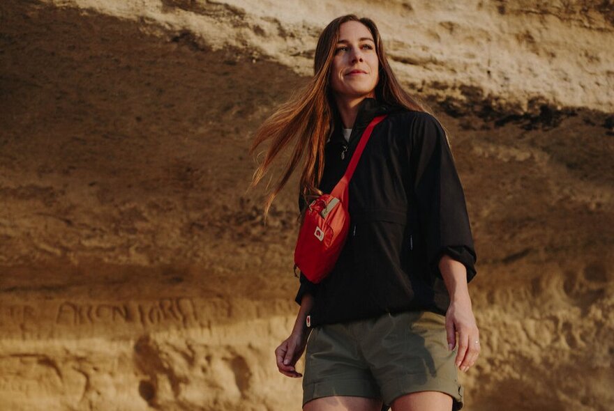 A woman in front of a natural rock formation wearing hiking gear and carrying a red Fjallraven sling bag.